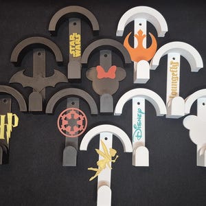Könnte beinhalten: Eine Sammlung dekorativer Wandhaken in verschiedenen Designs. Die Haken zeigen Themen wie Star Wars, Disney und Harry Potter. Farben sind Grau, Weiß, Orange und Gelb. Die Haken dienen zum Aufhängen von Gegenständen und verleihen eine dekorative Note.