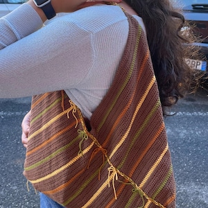 Pode incluir: Bolsa de ombro de crochê marrom com listras diagonais em tons de laranja, amarelo e verde. A bolsa tem formato triangular e é adornada com detalhes de fio amarrado nas bordas. A bolsa é usada no ombro.