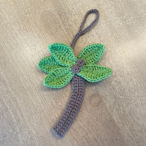 Puede incluir: Adorno de palmera hecho a mano con ganchillo. El árbol presenta una copa verde y verde claro, un tronco gris y un lazo para colgar. El adorno está hecho de hilo y está sobre un fondo de madera.
