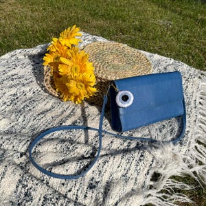 May include: A blue crossbody bag with a white and black eye charm, a woven basket, and yellow flowers are arranged on a white and grey blanket. The bag has a long strap and a flap closure.