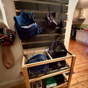 Puede incluir: Un organizador de entrada de madera con un tablero trasero teñido oscuro y un estante de madera natural. El tablero trasero tiene ganchos para colgar bolsos y sombreros, con los nombres "Charlotte" y "Tilly" escritos encima. El estante contiene zapatos y más bolsos.