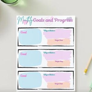 May include: A white planner with the text "Monthly Goals and Progress" in a script font. The planner has three sections labeled "Goal," "Why it Matters," and "Target Date." A pencil and a set of keys are next to the planner.