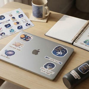 May include: A silver laptop decorated with space-themed stickers including astronauts and planets, surrounded by two mugs with star patterns, a sticker sheet, a spiral-bound notebook with space illustrations, and a black water bottle with a sticker.