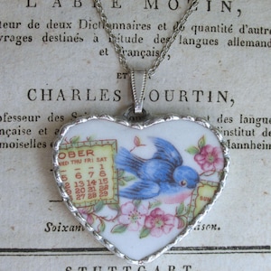 May include: A heart-shaped pendant on a silver chain featuring a white ceramic surface with a blue bird, pink flowers, and a partial calendar page showing the month of October with dates 1 through 29 and days of the week listed in black text.