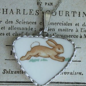 May include: A heart-shaped pendant necklace featuring a white ceramic center with a brown rabbit illustration. The pendant is encased in a silver-toned metal frame with a scalloped edge and hangs from a matching silver-toned metal chain link necklace.