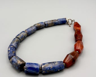 May include: A necklace featuring a combination of blue and red-orange gemstone beads. The blue beads are cylindrical with gold flecks, while the red-orange beads are faceted. The necklace has a silver clasp.