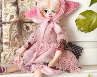May include: A handmade doll with a pink hooded outfit and a matching dress. The doll has a pale complexion, blonde hair, and detailed facial features. The outfit includes a pink bow and a polka-dot hood. The doll is sitting on a white surface.