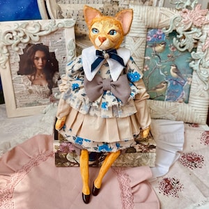 May include: A handcrafted cat doll with orange fur and blue eyes, wearing a floral dress with a white collar and a large gray bow. The doll is posed with its legs crossed, and it has brown shoes. The background includes framed artwork.
