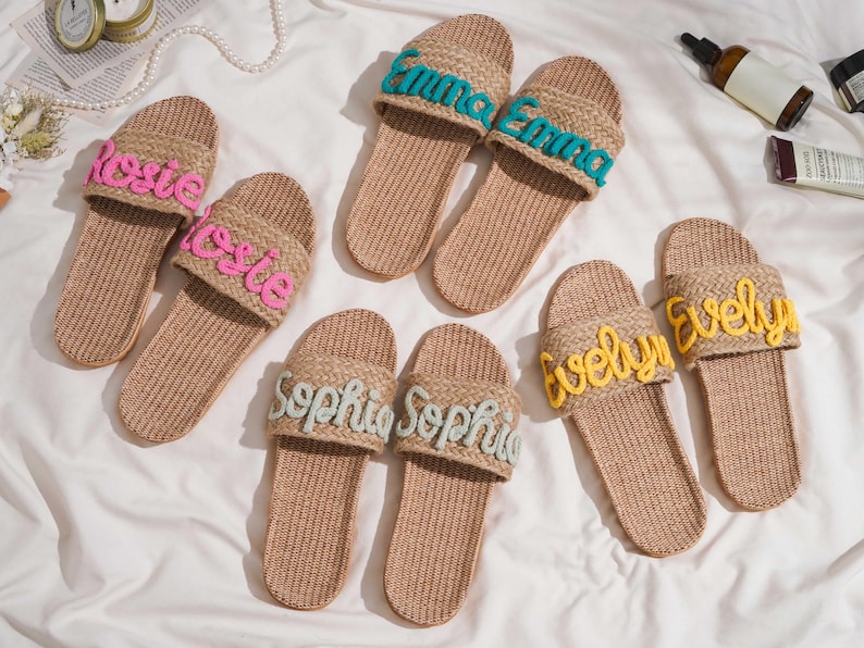 K&ouml;nnte beinhalten: Mehrere Paare gewebter Jute-Hausschuhe mit personalisierten Namen, die in verschiedenen Farben gestickt sind. Die Namen "Rosie", "Emma", "Sophia" und "Evelyn" sind in Rosa, T&uuml;rkis, Hellblau und Gelb dargestellt. Die Hausschuhe sind auf einer wei&szlig;en Oberfl&auml;che angeordnet.