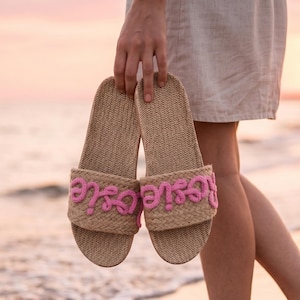 K&ouml;nnte beinhalten: Ein Paar gewebte beige Sandalen mit rosa Schreibschrift "Rosie" auf der Oberseite. Die Sandalen werden vor einer Strandszene bei Sonnenuntergang gehalten.