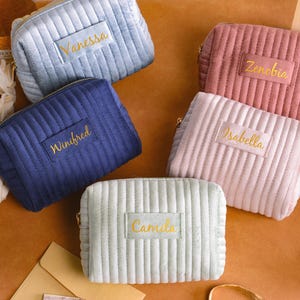 May include: Five rectangular velvet cosmetic bags with vertical channel quilting and gold zippers. Each bag features a rectangular patch with a name embroidered in gold thread: Vanessa, Zenobia, Winifred, Isabella, and Camila in various pastel colors.