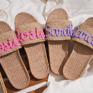 K&ouml;nnte beinhalten: Vier gewebte Raffia-Sandalen mit personalisierten Namen, die mit rosa und lila Faden gestickt sind. Die Namen "Rosie", "Sarah" und "Sarah" sind sichtbar. Die Sandalen haben eine nat&uuml;rliche beige Farbe und sind f&uuml;r den Freizeitgebrauch konzipiert.