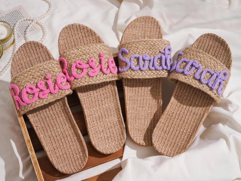K&ouml;nnte beinhalten: Vier gewebte Raffia-Sandalen mit personalisierten Namen, die mit rosa und lila Faden gestickt sind. Die Namen "Rosie", "Sarah" und "Sarah" sind sichtbar. Die Sandalen haben eine nat&uuml;rliche beige Farbe und sind f&uuml;r den Freizeitgebrauch konzipiert.