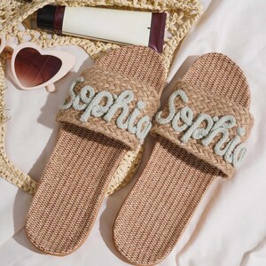 K&ouml;nnte beinhalten: Zwei beige gewebte Sandalen, jede mit dem Namen "Sophia" in hellblauer Schrift bestickt. Die Sandalen werden auf einer wei&szlig;en Oberfl&auml;che pr&auml;sentiert, mit einer gewebten Tasche, Sonnenbrille und Hautpflegeprodukten.