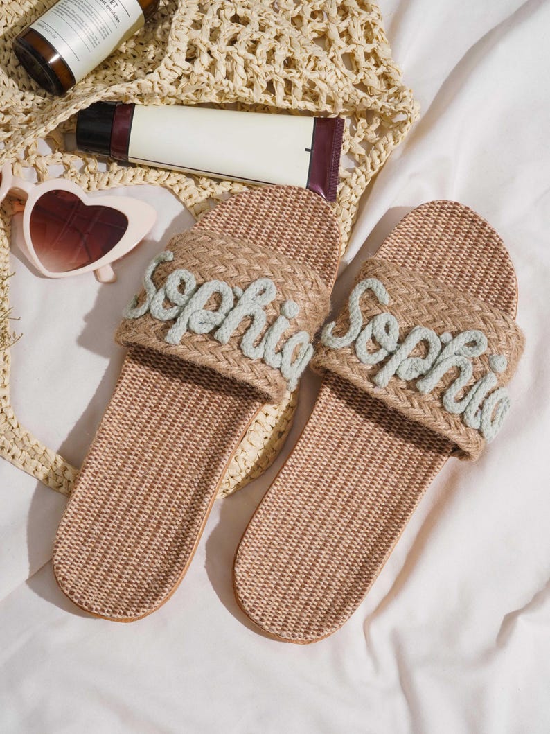 K&ouml;nnte beinhalten: Zwei beige gewebte Sandalen, jede mit dem Namen "Sophia" in hellblauer Schrift bestickt. Die Sandalen werden auf einer wei&szlig;en Oberfl&auml;che pr&auml;sentiert, mit einer gewebten Tasche, Sonnenbrille und Hautpflegeprodukten.