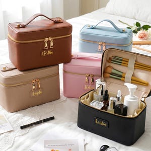 May include: A collection of five rectangular cosmetic cases in brown, light blue, beige, pink, and black. Each case features a top handle, gold-tone zippers, and a rectangular name tag with gold lettering. One black case is open, revealing makeup brushes and bottles.