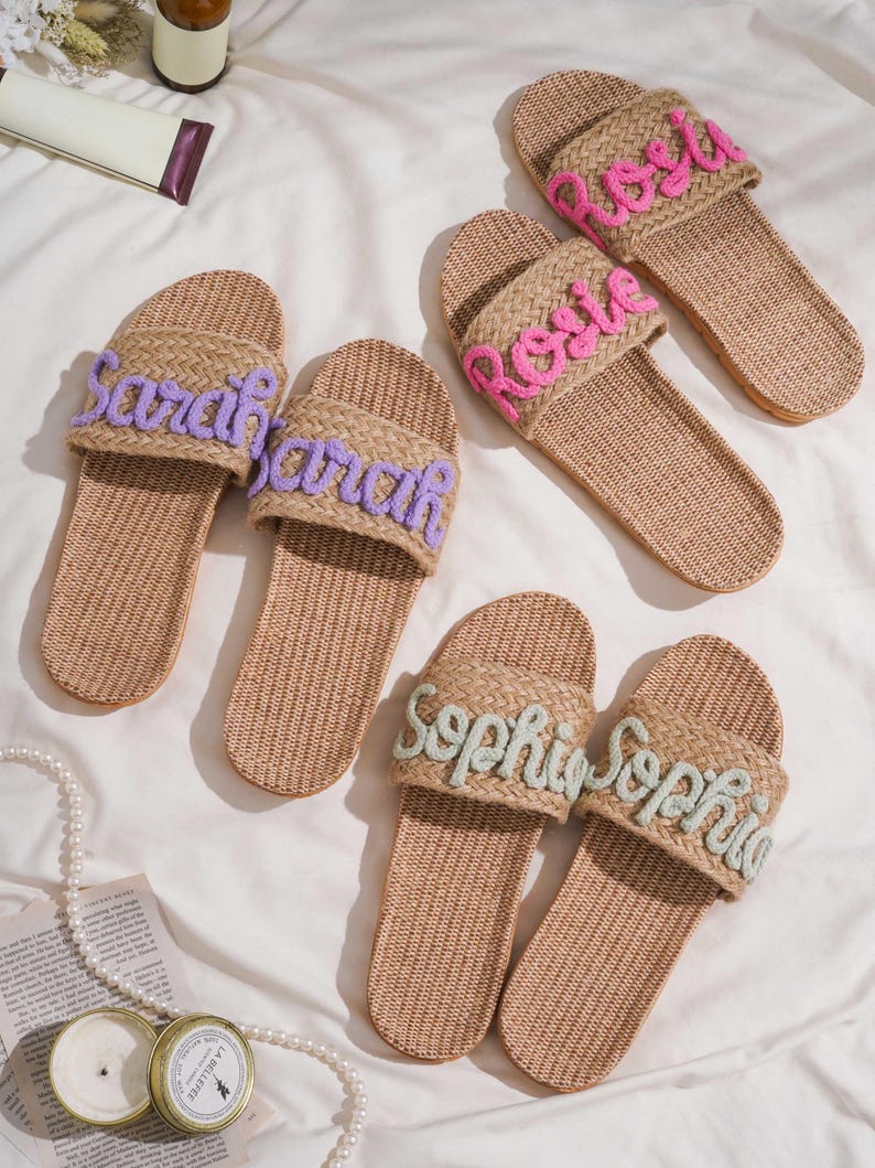 K&ouml;nnte beinhalten: Sechs Paar gewebte Sandalen mit gestickten Namen in Lila, Rosa und Hellgr&uuml;n. Die Namen "Sarah", "Rosie" und "Sophia" sind auf der Oberseite zu sehen. Die beigefarbenen Sandalen liegen auf einer wei&szlig;en Oberfl&auml;che.