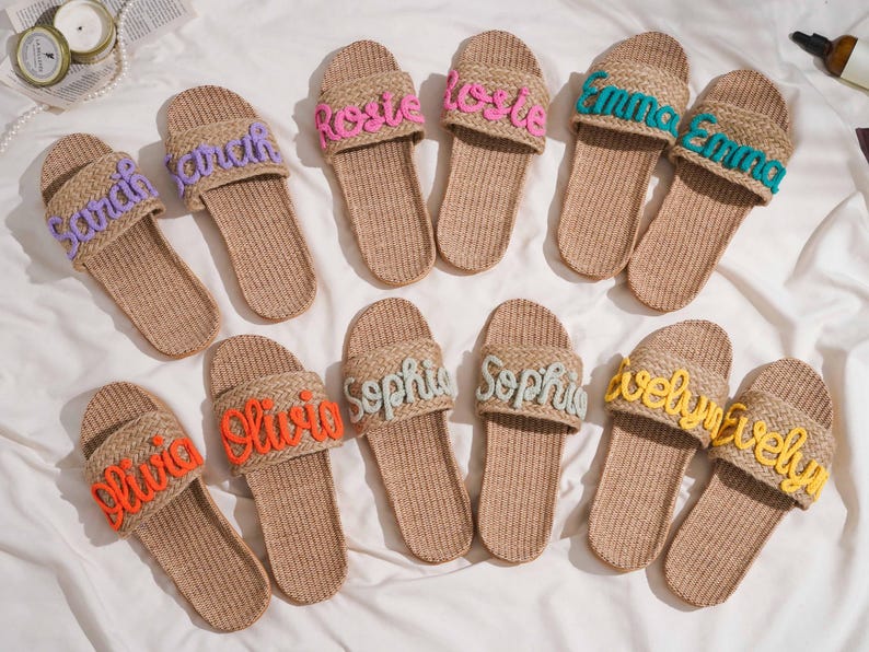 K&ouml;nnte beinhalten: Eine Auswahl an gewebten Jute-Hausschuhen mit farbenfrohen, gestickten Namen. Die Namen sind "Sarah", "Rosie", "Emma", "Olivia", "Sophie" und "Evelyn". Die Hausschuhe liegen auf einer wei&szlig;en Oberfl&auml;che.