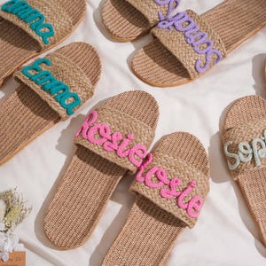 K&ouml;nnte beinhalten: Beige geflochtene Sandalen mit gestickten Namen in verschiedenen Farben. Die Sandalen haben ein strukturiertes Design und sind auf einer wei&szlig;en Oberfl&auml;che angeordnet. Die Namen sind "Emma", "Sarah", "Rosie" und "Sophie".