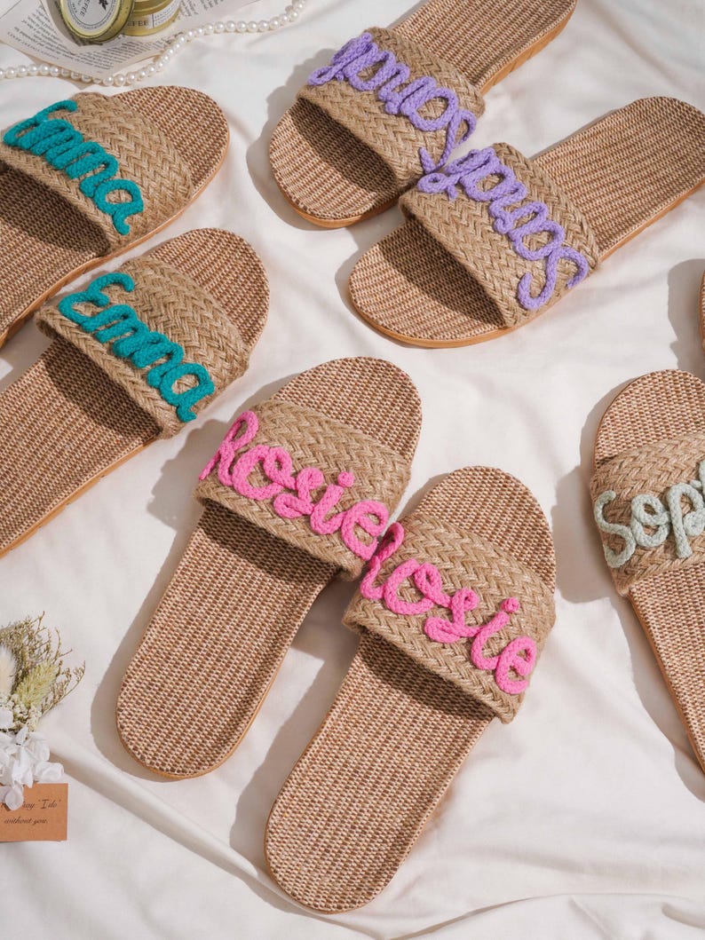 K&ouml;nnte beinhalten: Beige geflochtene Sandalen mit gestickten Namen in verschiedenen Farben. Die Sandalen haben ein strukturiertes Design und sind auf einer wei&szlig;en Oberfl&auml;che angeordnet. Die Namen sind "Emma", "Sarah", "Rosie" und "Sophie".
