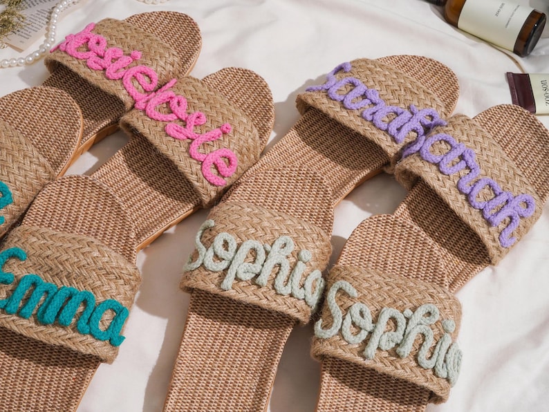 K&ouml;nnte beinhalten: Gewebte Raffia-Sandalen mit in verschiedenen Farben gestickten Namen. Die Namen sind "Emma", "Sophie", "Sarah" und "Rosie". Die Sandalen haben eine nat&uuml;rliche, neutrale Farbe. Personalisierung.