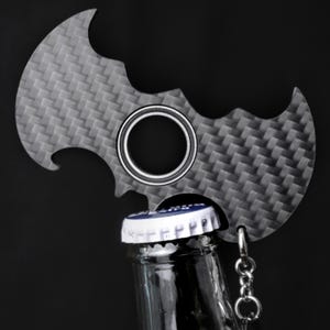 Könnte beinhalten: Ein grauer Flaschenöffner in Form eines Fledermaussymbols mit einem strukturierten Muster. Der Öffner hat eine kreisförmige Öffnung und ist an einem Flaschenverschluss befestigt. Eine kleine Kette mit einem Ring ist angebracht.