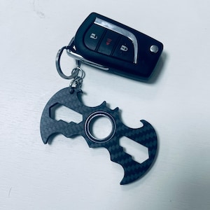 Könnte beinhalten: Ein schwarzer Autoschlüsselanhänger mit einem silbernen Schlüsselring, der an einem dunkelgrauen, fledermausförmigen Multi-Tool befestigt ist. Das Multi-Tool hat ein strukturiertes Karbonfasermuster und eine zentrale kreisförmige Öffnung.