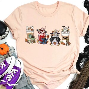 Könnte beinhalten: Hellrosa T-Shirt mit einem Halloween-Motiv. Das Design zeigt vier verschiedene Cartoon-Getränke, jedes mit einer Halloween-Figur, wie Baby Yoda, Stitch und Groot. Die Getränke sind mit Halloween-Elementen wie Kürbissen und Fledermäusen verziert.