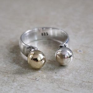 May include: A silver ring with two ball accents, one gold and one silver. The ring has a simple, open design. The metal is stamped with "925". The ring is displayed on a neutral surface.