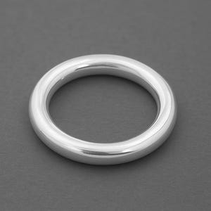 Könnte beinhalten: Ein massiver, polierter Silberring mit glattem, abgerundetem Design. Der Ring hat eine kreisförmige Form und eine helle, reflektierende Oberfläche. Der Schmuck ist vor einem dunkelgrauen Hintergrund platziert.