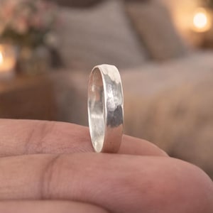 以下が含まれることがあります： ハンマー加工されたテクスチャのシルバーリングが指で支えられています。リングはシンプルなバンドデザインです。背景はぼやけており、柔らかな照明のある寝室の様子が写っています。