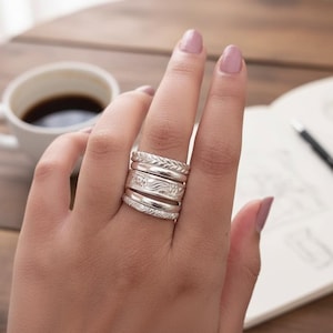 May include: A hand wearing multiple silver rings with various designs, including braided, floral, and textured patterns. The rings are stacked on the finger, showcasing a collection of jewelry. A cup of coffee and a notebook are in the background.