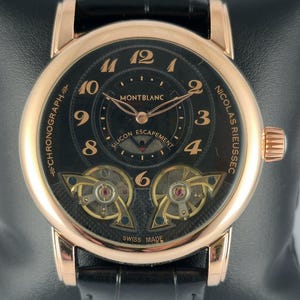 Peut inclure: Une montre de luxe avec un cadran noir et des détails dorés. La montre porte l'inscription "MONTBLANC" et "SWISS MADE". Elle est dotée d'un bracelet en cuir noir et d'un boîtier doré. La montre est présentée sur une surface noire.