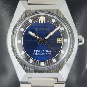 Puede incluir: Un reloj automático King Seiko plateado con esfera azul y visualización de fecha. El reloj tiene una caja rectangular y una correa de acero inoxidable. La esfera presenta marcadores y agujas blancas, con el texto "King Seiko Automatic 3 Days".