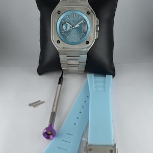 May include: A stainless steel watch with a square face and a light blue dial. The watch has a matching stainless steel band. Also included are a light blue watch strap, a screwdriver, and two small silver pins. The watch is displayed on a black pillow.