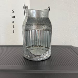 May include: A small, galvanized metal milk can-shaped container with a handle and vertical ribbed design. The container has a rustic, industrial aesthetic and is suitable for home decor or storage. The word "small" is written vertically on the side.
