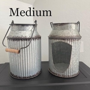 May include: Two galvanized metal milk cans with a rustic, distressed finish. One has a wooden handle and a hinged lid. The other has a large opening in the front. The word "Medium" is above the cans.