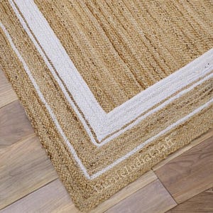 May include: A rectangular jute rug with a natural tan color and a white border. The rug has a woven texture and is placed on a wooden floor. The rug's dimensions are not visible in the image.