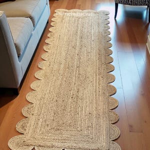 Op de afbeelding: Een rechthoekige jute loper met een geschulpte rand. Het tapijt heeft een natuurlijke beige kleur en een geweven textuur. Het tapijt ligt op een hardhouten vloer. De tekst "DurmisIndiaCraft" is zichtbaar.