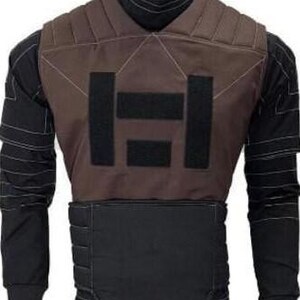 May include: A brown tactical vest featuring a black geometric design on the front. The vest is worn over a black long-sleeved shirt with white stitching. The vest appears to be made of a durable material, possibly ballistic.