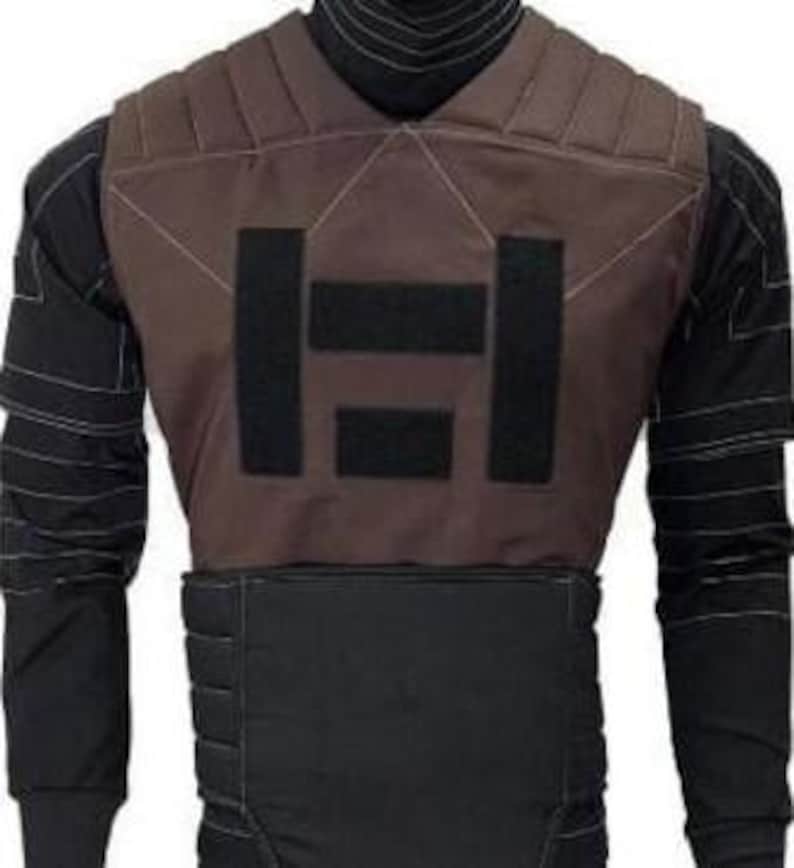 May include: A brown tactical vest featuring a black geometric design on the front. The vest is worn over a black long-sleeved shirt with white stitching. The vest appears to be made of a durable material, possibly ballistic.