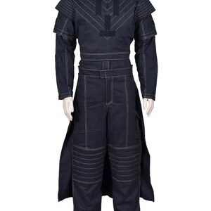 May include: A dark navy blue long-sleeved jumpsuit featuring white contrast stitching throughout. The garment includes a structured bodice with shoulder panels, a belted waist, and pleated knee sections. A long, open-sided skirt overlay extends from the waist down to the hem, creating a layered appearance.
