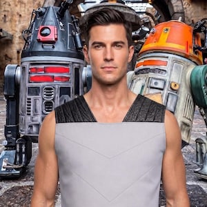 May include: A sleeveless light gray tunic featuring black textured shoulder panels and a subtle V-shaped stitched pattern on the front. The garment is worn by a person standing between a black and red robotic figure and an orange and white droid.