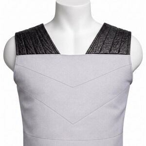 May include: A sleeveless vest featuring a light gray fabric body with a V-shaped stitched chevron pattern across the chest. The shoulders are accented with wide, black textured panels that have vertical stitching details along the top.