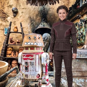 May include: A person wearing a dark brown long-sleeved tunic with a high collar and matching trousers, standing next to a weathered, cylindrical droid with a white, red, and tan finish, featuring a painted jagged tooth design on its dome.