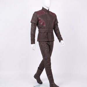 May include: A brown long-sleeved tunic and matching trousers set featuring dark maroon shoulder panels, a high collar, and a waist belt. The garment includes white stitched geometric patterns on the sleeves and linear detailing throughout the fabric.