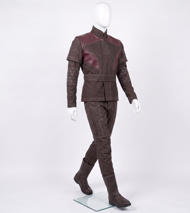 May include: A brown long-sleeved tunic and matching trousers set featuring dark maroon shoulder panels, a high collar, and a waist belt. The garment includes white stitched geometric patterns on the sleeves and linear detailing throughout the fabric.