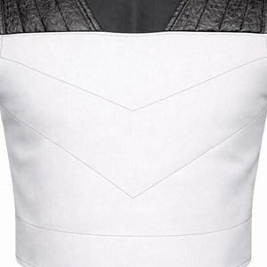 May include: A white vest featuring a V-shaped stitched seam across the chest and black textured leather panels on the shoulders. The garment has a clean, structured design with a horizontal band at the bottom and a high neckline.