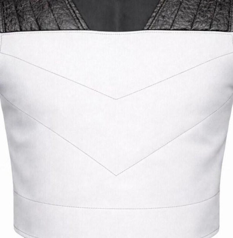 May include: A white vest featuring a V-shaped stitched seam across the chest and black textured leather panels on the shoulders. The garment has a clean, structured design with a horizontal band at the bottom and a high neckline.