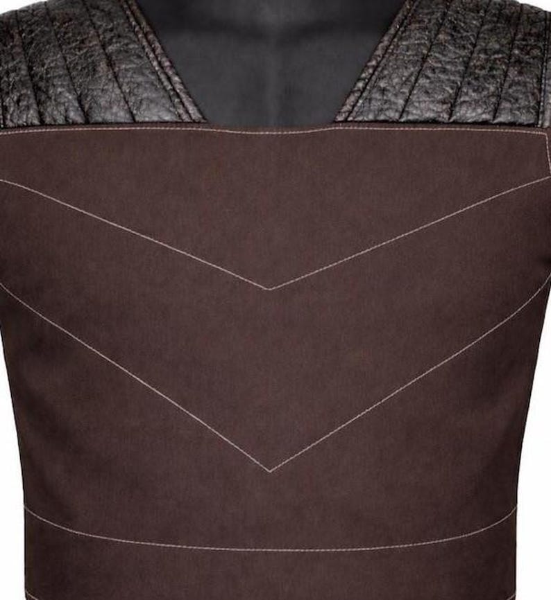 May include: A dark brown vest featuring a smooth matte finish on the main body with contrasting black textured shoulder panels. The garment includes prominent white topstitching that forms a large chevron pattern across the chest area.